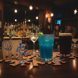 Puede incluir: Una escena de bar con un cóctel azul, un martini y una cerveza oscura. Posavasos de madera con un oso teñido anudado y un logotipo de Stealie en primer plano. El bar tiene un acabado de madera oscura y está tenuemente iluminado.