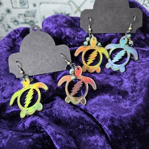 Könnte beinhalten: Ein Paar regenbogenfarbene, tie-dye, schildkrötenförmige Ohrringe mit einem Blitz-Design. Die Ohrringe sind aus Holz und haben silberne Haken.