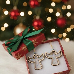 May include: A pair of angel-shaped earrings with glitter filling, displayed in a red gift box with a green ribbon. The box is decorated with gold snowflakes. The background features a Christmas tree with ornaments.