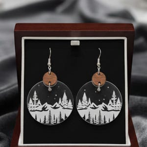 Puede incluir: Pendientes redondos con un diseño de montaña y bosque. Los pendientes transparentes tienen un gráfico blanco y un pequeño acento de madera. Se presentan en una caja de joyería de madera oscura.