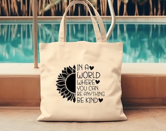 Peut inclure: Un sac fourre-tout beige avec du texte noir et un graphique de tournesol. Le texte dit "Dans un monde où vous pouvez être tout ce que vous voulez, soyez gentil".