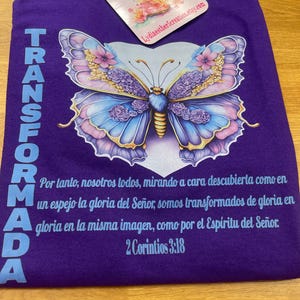 May include: Purple t-shirt featuring a large butterfly design in shades of blue, pink, and purple. The word "TRANSFORMADA" is printed vertically on the left side. The shirt also includes text from 2 Corinthians 3:18 in Spanish.
