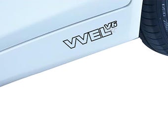 Peut inclure: Un panneau latéral de voiture blanc avec un autocollant au contour noir portant le texte VVEL V6. L'autocollant est positionné horizontalement le long du panneau inférieur, près du pneu arrière, qui montre un pneu en caoutchouc noir avec une jante en alliage argenté.