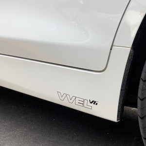 May include: A white car with a black "VVVL V6" sticker on the side.