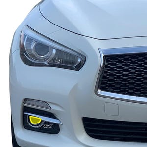 May include: The front passenger side of a white car featuring a bright yellow fog light tint decal applied to the circular light housing. The decal includes the small white text TINT DECAL printed on the lower edge of the black trim surround.
