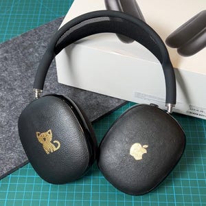 Peut inclure: Casque audio noir avec un motif de chat doré sur une oreillette et le logo Apple doré sur l'autre. Le casque a un bandeau en maille noire. Le casque est sur un tapis de coupe vert.