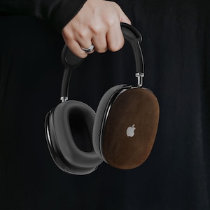 Könnte beinhalten: Schwarzer Over-Ear-Kopfhörer mit braunen Leder-Ohrmuscheln und einem silbernen Apple-Logo. Der Kopfhörer hat ein schwarzes Kopfband und Ohrpolster. Der Kopfhörer wird von einer Person gehalten, die ein schwarzes Oberteil trägt.