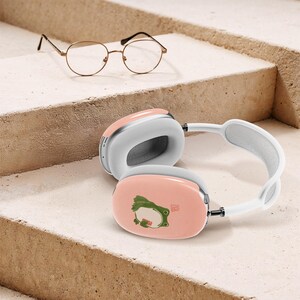 Könnte beinhalten: Rosa-weiße Over-Ear-Kopfhörer mit einem Cartoon-Frosch-Design. Die Kopfhörer werden mit einer goldgerahmten Brille kombiniert. Die Kopfhörer haben ein weißes Kopfband und Ohrmuscheln mit hellrosa Außenbereich.