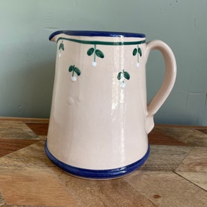 May include: A cream-colored ceramic pitcher with a blue and green floral design. The pitcher has a wide mouth and a handle.