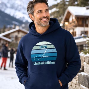 May include: A navy blue hooded sweatshirt featuring a graphic of a sunset with horizontal stripes in shades of blue and green. The graphic includes the text 1976, the name Arne in a script font, and the words Limited Edition printed below.