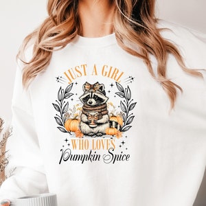 Könnte beinhalten: Weißes Sweatshirt mit einem Aufdruck, der einen Waschbären mit Schal und Tasse, umgeben von Kürbissen und Herbstlaub, zeigt. Der Text lautet: "JUST A GIRL WHO LOVES Pumpkin Spice".
