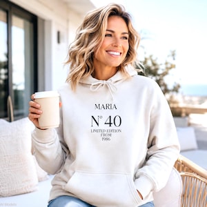 Könnte beinhalten: Ein cremefarbener Kapuzenpullover mit einer Kängurutasche vorne und einer Kapuze mit Kordelzug. Auf der Brust befindet sich ein schwarzer Schriftzug mit dem Text MARIA, N° 40, LIMITED EDITION und FROM 1986 in einer Kombination aus serifenloser Schrift.