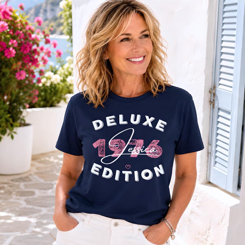 May include: A navy blue short-sleeve t-shirt featuring the white text DELUXE EDITION with the pink textured numbers 1976 and the name Jessica in white script in the center, accompanied by a small pink heart icon below the text.