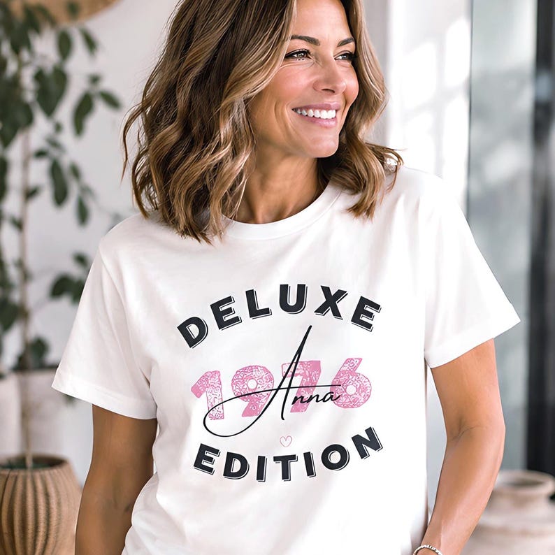May include: A white short-sleeved t-shirt featuring the black text DELUXE EDITION in a bold font. Centered between these words is the year 1976 in a textured pink pattern, with the name Anna written in black script and a small pink heart icon.
