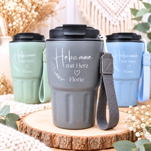 Könnte beinhalten: Drei isolierte Thermobecher in Salbeigrün, Dunkelgrau und Hellblau. Jeder Becher hat einen schwarzen Deckel, eine passende Silikonschlaufe und den weißen Schriftzug Hebamme mit Herz, ein Herzsymbol sowie den Namen Florie mit Blumenmotiv.