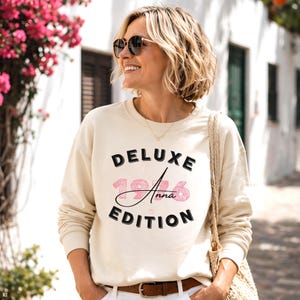 Könnte beinhalten: Ein cremefarbenes Langarm-Sweatshirt mit Rundhalsausschnitt, das auf der Vorderseite den schwarzen Schriftzug DELUXE EDITION sowie die rosafarben gemusterten Zahlen 1976 mit dem Namen Anna in schwarzer Schreibschrift zeigt.