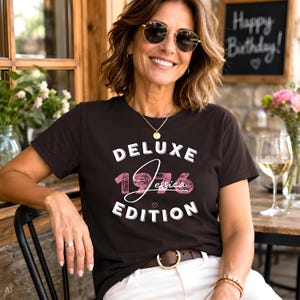 May include: A dark brown short-sleeved t-shirt featuring the white text DELUXE EDITION and the pink textured number 1976 with the name Jessica written in white script across the center, accompanied by a small pink heart icon below the name.