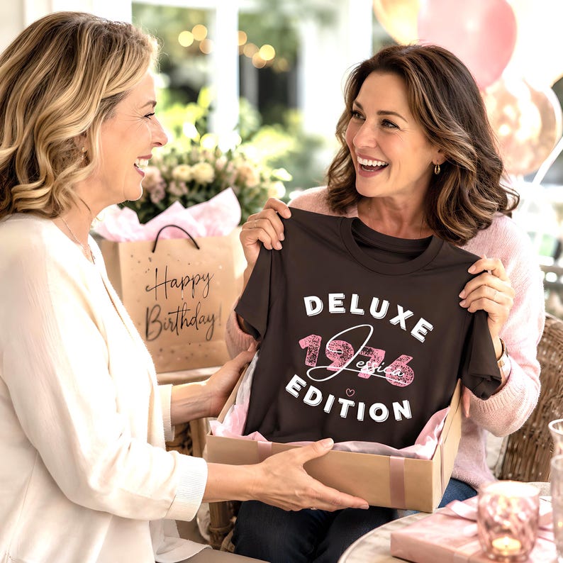 May include: A dark brown short-sleeved t-shirt featuring the white text "DELUXE EDITION" and the pink glittery number "1976" with the name "Jessica" written in white script across the center, all displayed inside a cardboard gift box.