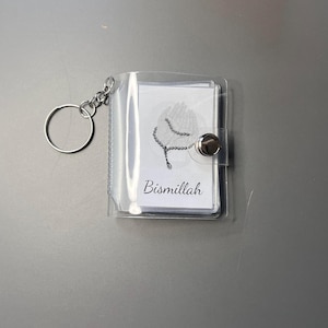 Può includere: Un portachiavi a forma di mini libro in plastica trasparente con un anello in metallo. La copertina presenta un inserto bianco con un'illustrazione in bianco e nero di mani che tengono grani da preghiera e la parola Bismillah in carattere corsivo, chiusa da un bottone a pressione.