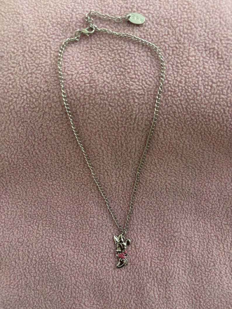 May include: A silver-toned necklace with a Minnie Mouse pendant. The pendant features Minnie's iconic bow with pink accents. The chain is delicate and hangs on a pink background. A small, round charm is near the clasp.