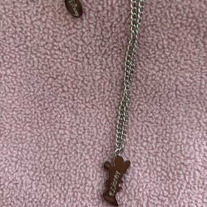 May include: Silver-toned necklace with a Disney pendant. The pendant is in the shape of a character and has the Disney logo. The necklace is on a pink textured background.