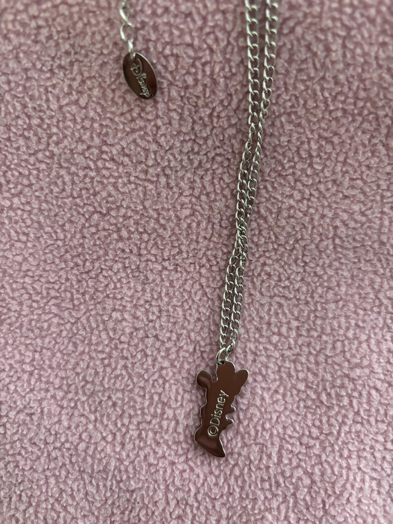 May include: Silver-toned necklace with a Disney pendant. The pendant is in the shape of a character and has the Disney logo. The necklace is on a pink textured background.