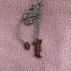 May include: A silver-toned necklace with a pendant in the shape of a cartoon character. The pendant is brown and features the text "&copy;Disney". The necklace is on a pink textured surface.