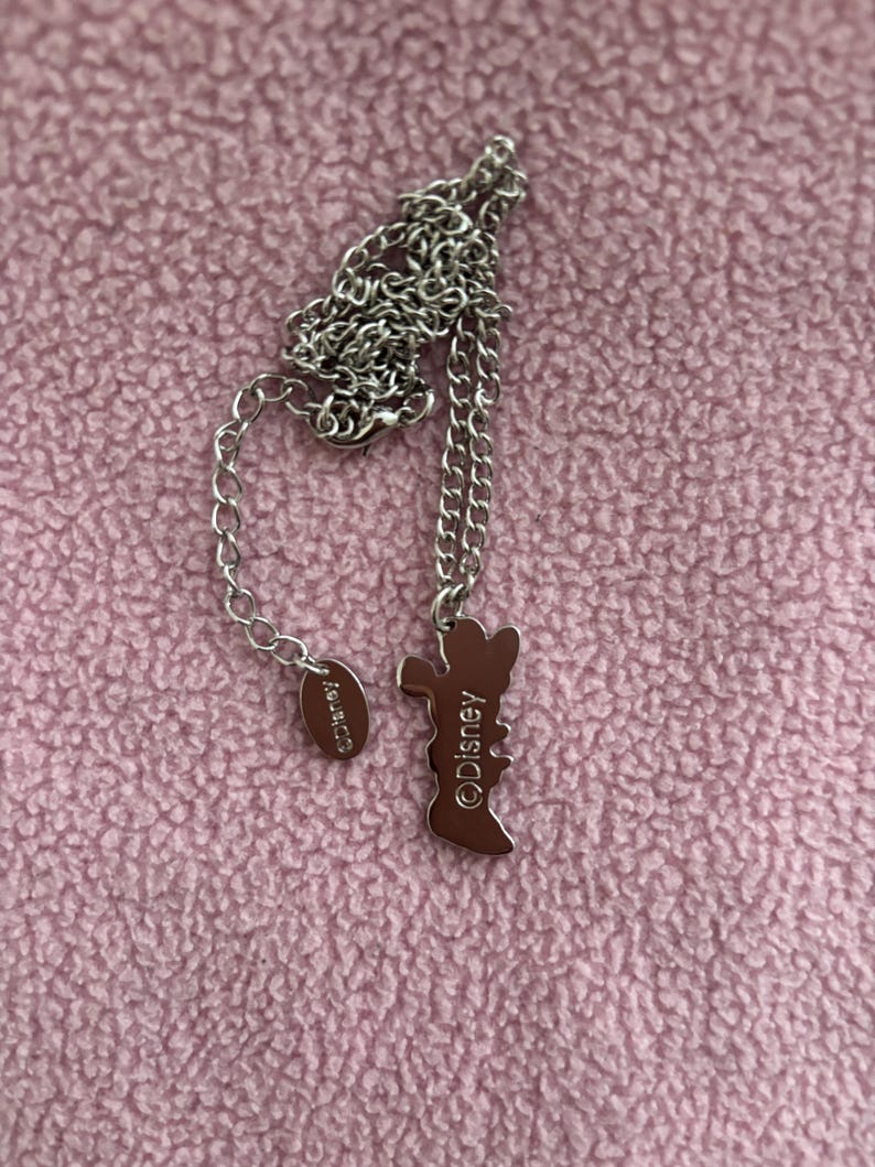 May include: A silver-toned necklace with a pendant in the shape of a cartoon character. The pendant is brown and features the text "&copy;Disney". The necklace is on a pink textured surface.