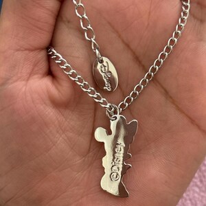 May include: A silver-colored necklace featuring a Mickey Mouse pendant. The pendant has the Disney logo engraved on it. The necklace is held in a hand.
