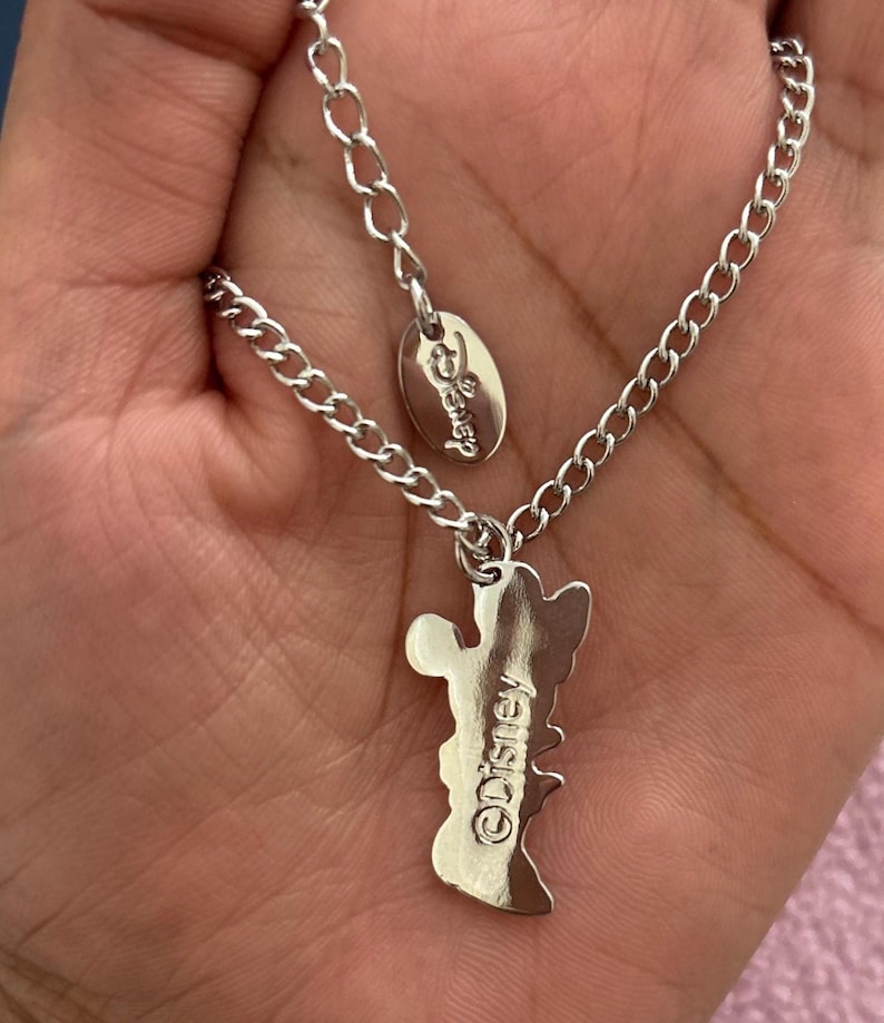 May include: A silver-colored necklace featuring a Mickey Mouse pendant. The pendant has the Disney logo engraved on it. The necklace is held in a hand.