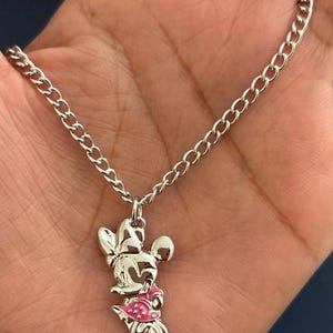 May include: Silver-toned necklace featuring a Minnie Mouse pendant. The pendant has a pink dress and bow, with a chain link necklace. The necklace is held in a hand.