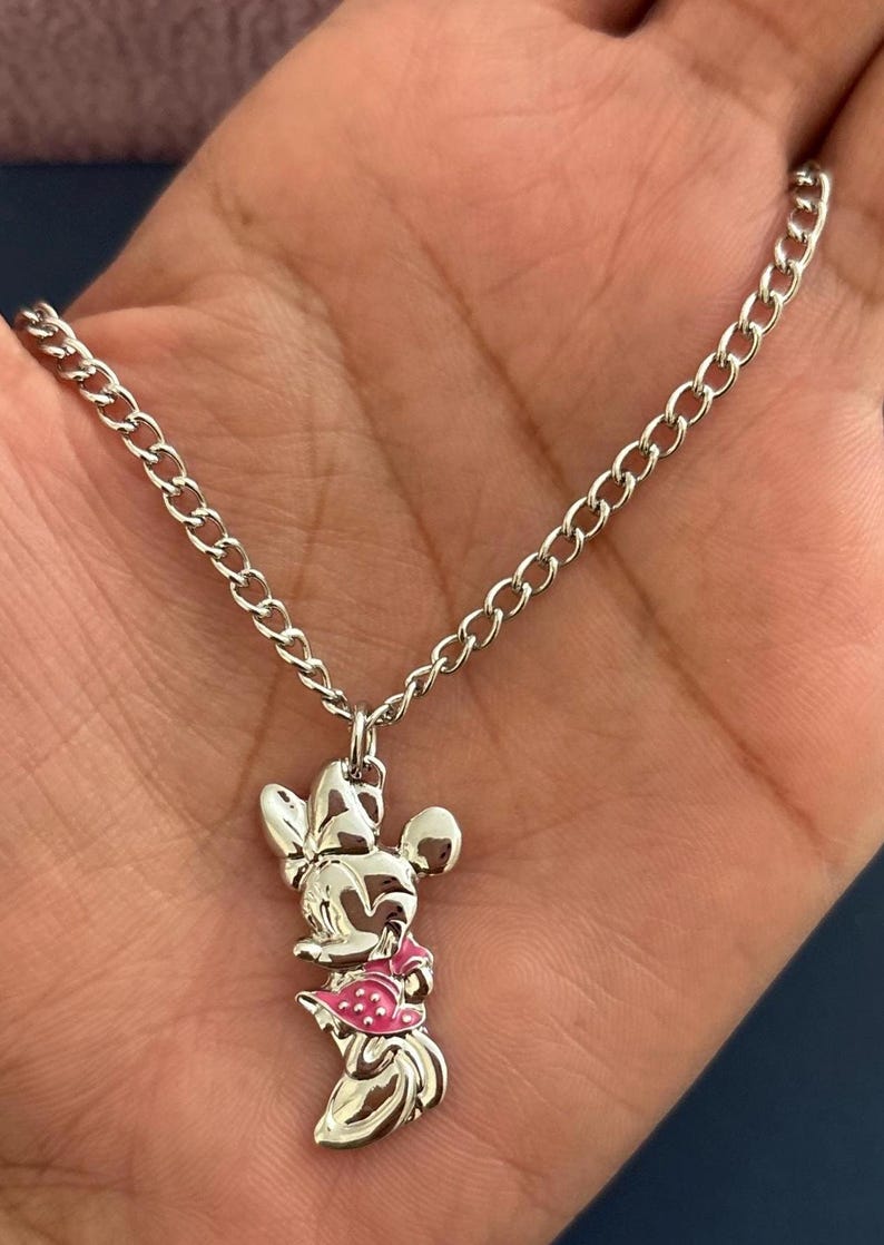 May include: Silver-toned necklace featuring a Minnie Mouse pendant. The pendant has a pink dress and bow, with a chain link necklace. The necklace is held in a hand.
