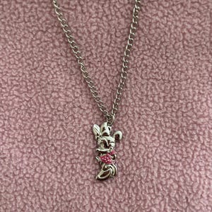 May include: Silver-toned necklace featuring a pendant of a cartoon character with a pink bow and dress. The pendant hangs from a chain against a pink textured background. The necklace is a piece of jewelry.
