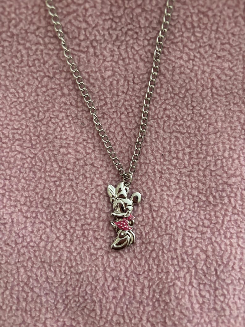 May include: Silver-toned necklace featuring a pendant of a cartoon character with a pink bow and dress. The pendant hangs from a chain against a pink textured background. The necklace is a piece of jewelry.