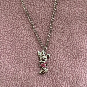 May include: Silver-toned necklace featuring a Minnie Mouse pendant with a pink bow and dress. The pendant is suspended from a delicate chain. The background is a textured, light pink fabric.