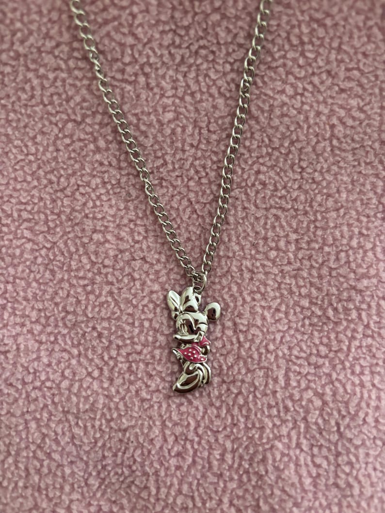 May include: Silver-toned necklace featuring a Minnie Mouse pendant with a pink bow and dress. The pendant is suspended from a delicate chain. The background is a textured, light pink fabric.