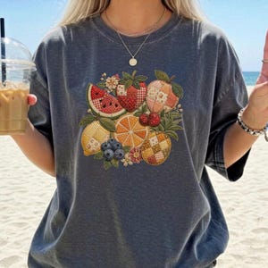 Puede incluir: Camiseta gris oscuro con un diseño de frutas colorido. El diseño incluye una sandía, fresas, una manzana, un limón, una naranja, arándanos y una piña. La fruta está dispuesta en un patrón decorativo con acentos florales.