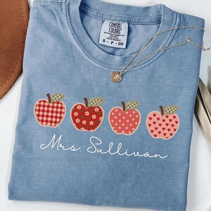 May include: A blue crew neck t-shirt featuring a graphic of four apples with different patterns including red gingham, floral, polka dot, and heart prints. Below the apples, the text Mrs. Sullivan is printed in white script. A gold necklace is visible.