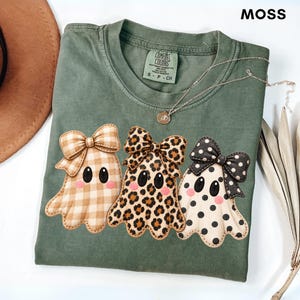 May include: A moss green short-sleeve t-shirt featuring a graphic of three ghosts wearing bows. The left ghost has a tan gingham pattern, the middle ghost has a leopard print, and the right ghost has black polka dots. The shirt has a Comfort Colors label.