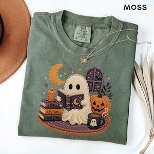 Może przedstawiać: Mchowo-zielony t-shirt z wyhaftowanym wzorem ducha czytającego książkę, otoczonego półksiężycem, świecą, stosem książek z napisami Spells, Potions i Ghost Stories, dynią oraz kubkiem z ikoną ducha.