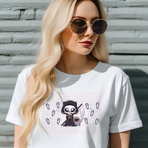May include: White t-shirt featuring a graphic of a cartoon Grim Reaper holding a small coffin. The graphic is surrounded by small white bones and coffins on a pink and white grid pattern. The Grim Reaper is wearing a black hooded robe and has a skull face. The coffin has the word "RIP" on it.