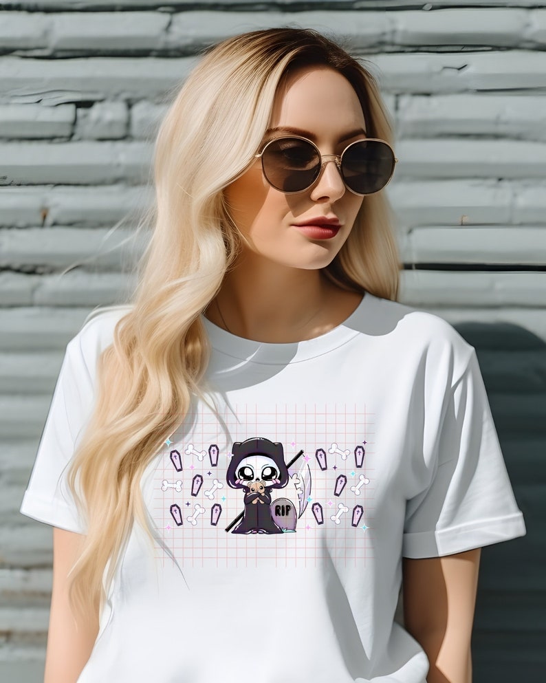 May include: White t-shirt featuring a graphic of a cartoon Grim Reaper holding a small coffin. The graphic is surrounded by small white bones and coffins on a pink and white grid pattern. The Grim Reaper is wearing a black hooded robe and has a skull face. The coffin has the word "RIP" on it.