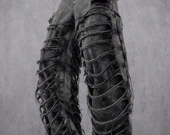 May include: Distressed black denim jeans with large rips and frayed edges. The jeans have a wide-leg fit and are styled with black boots. The design features multiple horizontal and vertical cuts, creating a unique, edgy look.