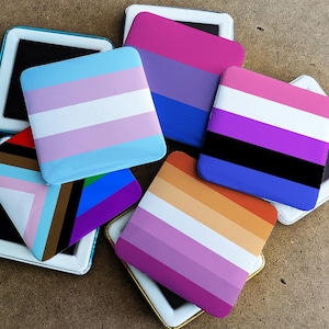 Op de afbeelding: Een verzameling vierkante items, elk met een ander LGBTQ+ pride vlagontwerp. De vlaggen omvatten transgender, biseksueel, non-binair, lesbisch en progress pride vlaggen. De items zijn geplaatst tegen een neutrale achtergrond.