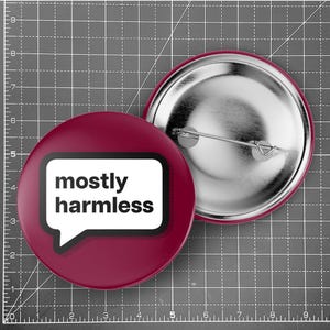 Könnte beinhalten: Zwei runde Ansteckbuttons. Einer ist weinrot mit den Worten "mostly harmless" in einer weißen Sprechblase. Der andere Button ist silberfarben mit einer Nadel. Sie liegen auf einer grauen Rasterfläche.