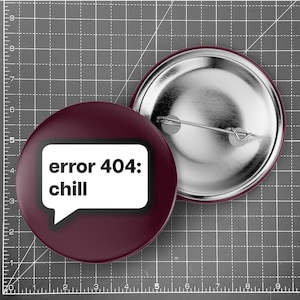 Könnte beinhalten: Zwei runde, weinrote Ansteckbuttons. Einer zeigt den Text "error 404: chill" in einer weißen Sprechblase. Der andere Button ist silberfarben und hat eine Nadel auf der Rückseite. Die Buttons liegen auf einem grauen Raster.