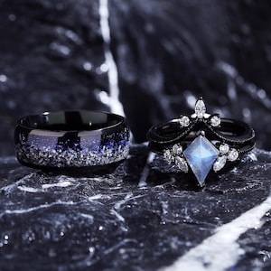 May include: A black wedding band with a blue and white galaxy design and a black engagement ring with a large, clear, faceted gemstone and smaller clear gemstones.