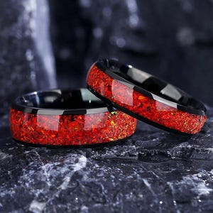 Könnte beinhalten: Zwei schwarze Ringe mit einem lebendigen rot-orangen, gesprenkelten Inlay. Die Ringe sind vor einem dunklen, strukturierten Hintergrund platziert, der den Kontrast zwischen den schwarzen Bändern und dem feurigen Inlay hervorhebt.