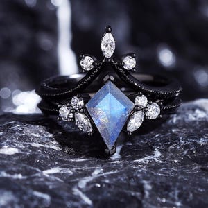 May include: A black ring with a large, blue, faceted gemstone surrounded by smaller, clear gemstones. The ring has a delicate, crown-like design.