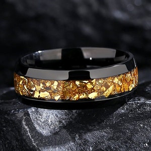 May include: A black ring with a gold inlay. The inlay is made of small, irregular pieces of gold that are scattered across the band.